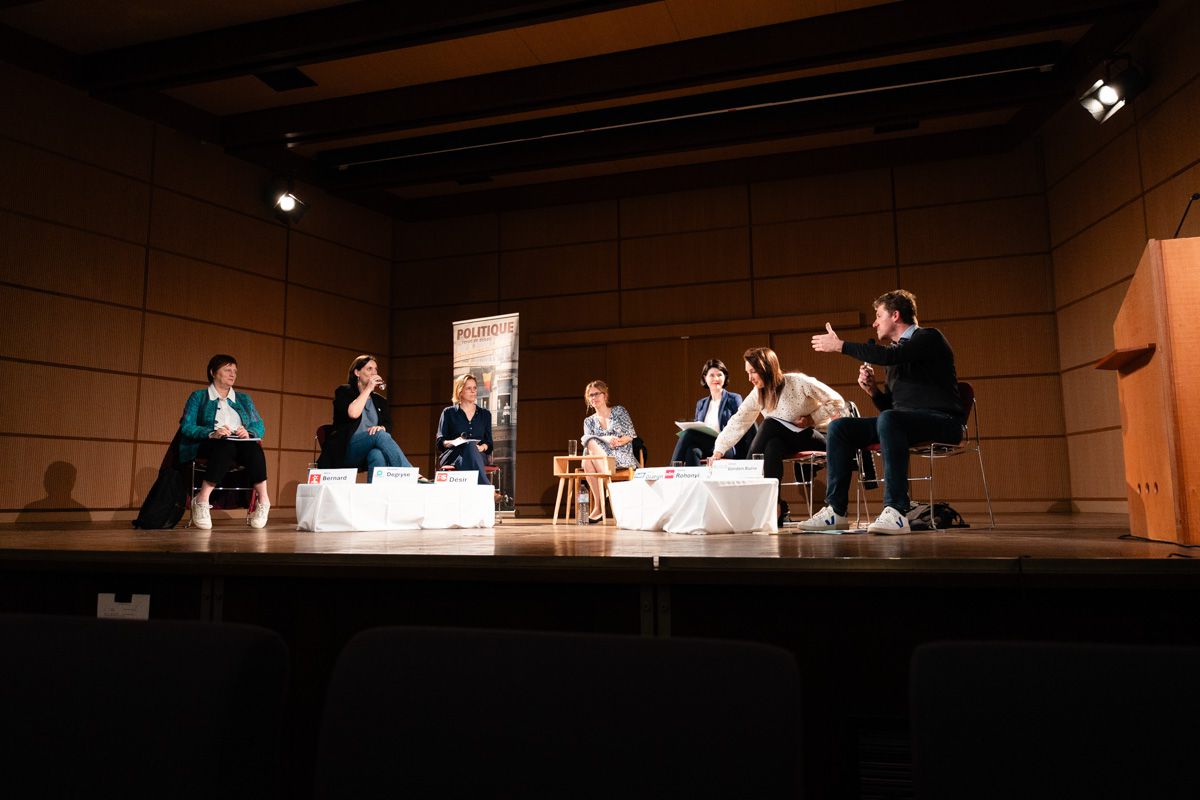 A panel of seven on a theatre stage with wood-panelled walls; a man on the right speaks with an arm stretched out, seen from the dark seats.