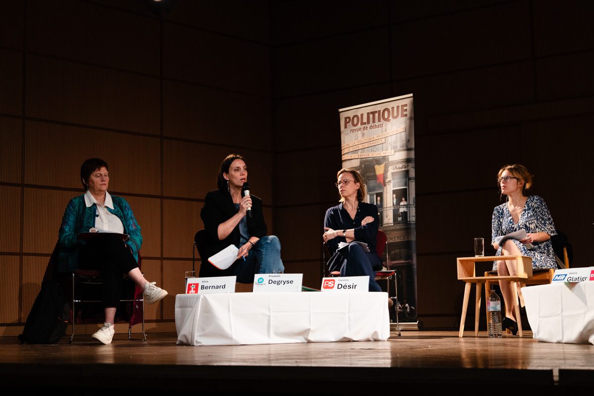 Four women seated on stage behind name cards; one speaks into a microphone, the others listen.