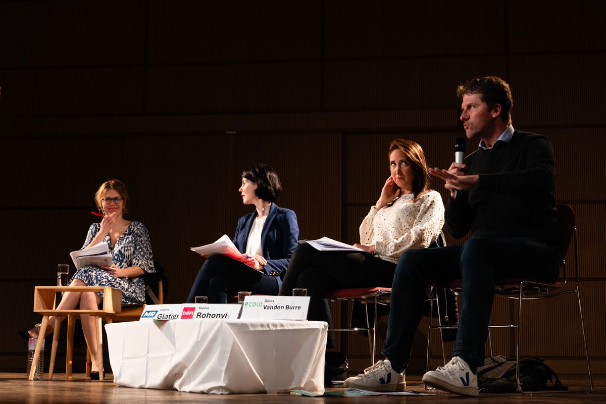 A man speaks into a microphone, hand raised; three women seated beside him on stage turn toward him.