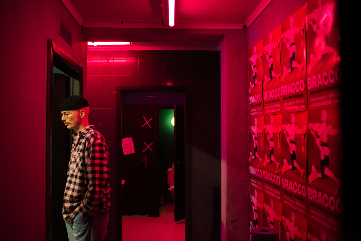 A man in a checked shirt and cap standing in a red-lit corridor next to posters.