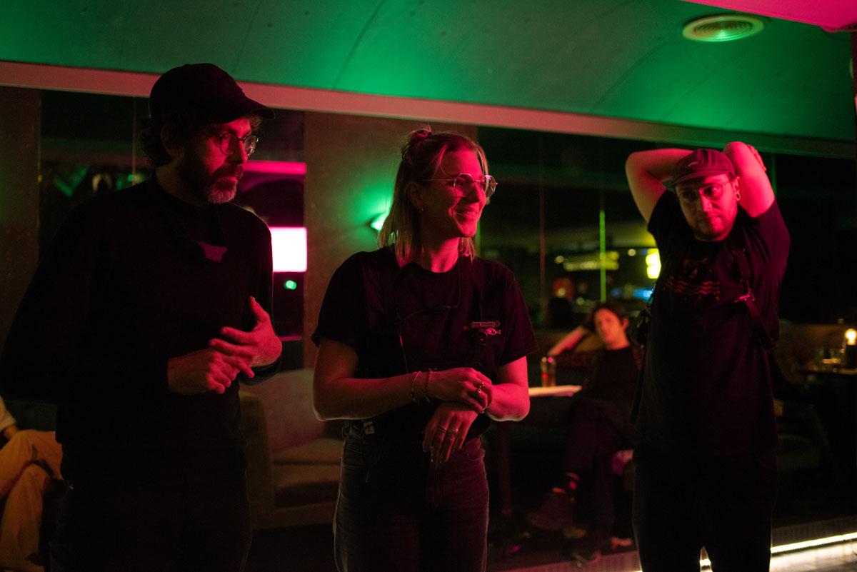 Three people standing in a bar lit in green, one with hands clasped, another with an arm raised.