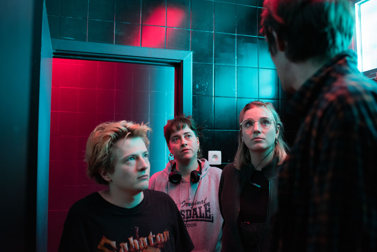 Four people looking out of frame, in front of a red door in a blue-lit tiled set.
