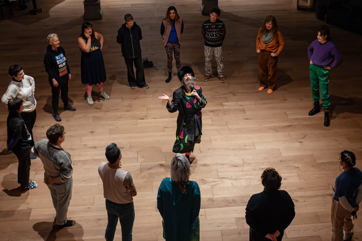 View from above: a performer at the centre of a circle of spectators on a light wooden floor.