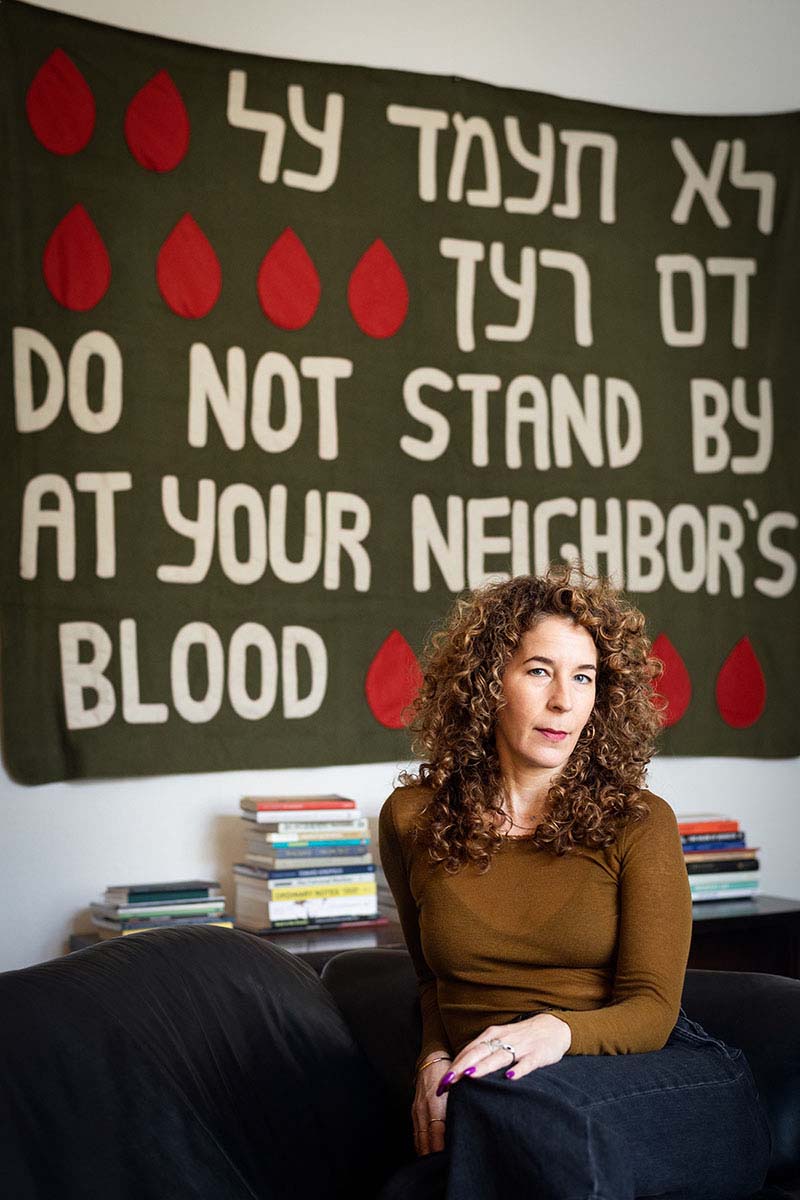 Portrait of a woman with curly hair seated on a sofa in front of a banner embroidered with drops of blood.