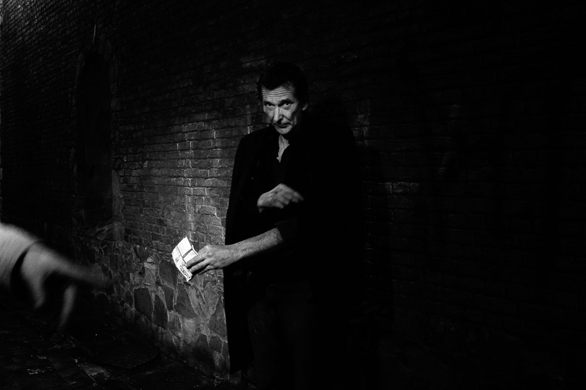 A man against a brick wall holds a banknote, a hand reaching into the frame.