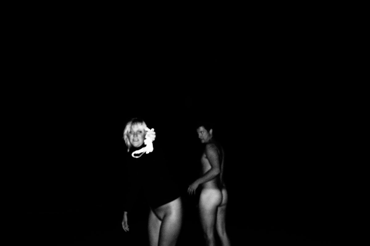 At night on a beach, a woman holds out her knickers toward the camera; a naked man is seen from behind.