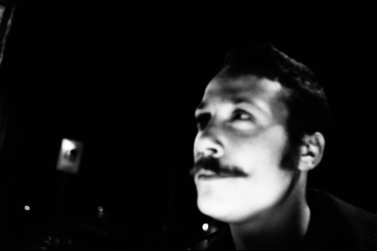 A man with a moustache looks up toward a light outside the frame.